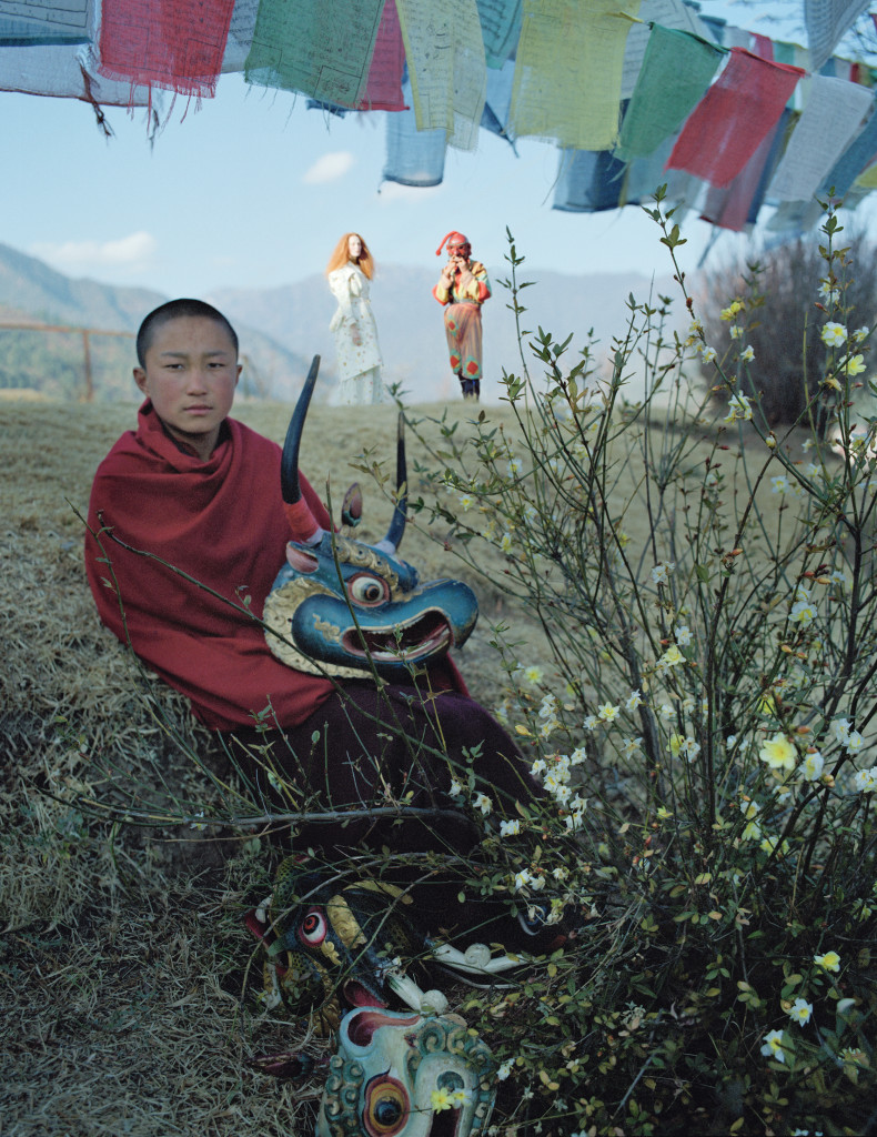 팀푸의 수도원 벽에 걸리게 될 초상화를 위해 포즈를 취한 동자승. 언덕 위의 카렌은 풍요로움과 다산의 상징인 앗사라(Atsara)의 탈을 쓴 스님과 대화를 나누고 있다. 실크 코트와 드레스는 로샤(Rochas).
